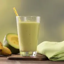 Fotografia em tons de verde e amarelo de uma bancada de madeira com um paninho verde e um apoio de copo de madeira, sobre ele um copo de vidro com a vitamina de abacate e um canudo amarelo. Ao fundo três bananas e meio abacate.