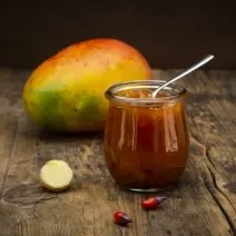 Fotografia de um chutney de manga e laranja temperado dentro de um recipiente de vidro pequeno, como se fosse um copo. Apoiado no chutney, uma colher de chá, e ao fundo, uma manga inteira com casca, sobre uma mesa de madeira.