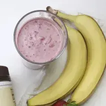 Fotografia em tons de rosa em uma mesa branca, uma toalha branca, duas bananas e um copo de vidro alto com o smoothie de morango com banana e pasta de amendoim dentro dele.