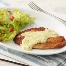 Fotografia em tons de azul em uma bancada de madeira clara com um pano listrado azul claro e um prato oval branco com o filé de pescada com molho de limão e mostrada e salada de alface com tomates.