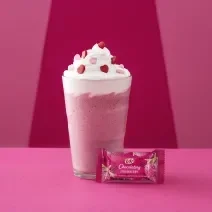 Foto da receita de Frappé. Observa-se um fundo rosa com um copo alto decorado de chantily e confeitos de coração com um kit kat de morango na frente.