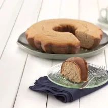 Fotografia em tons de verde em uma bancada de madeira branca, um paninho verde, um prato redondo grande com o bolo de amêndoas e aveia com um pedaço cortado e um pratinho pequeno com a fatia do bolo.