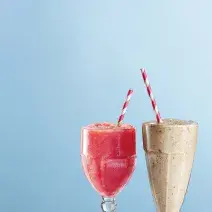 Fotografia em tons de azul e roxo vista de frente. Dois copos transparentes um com uma bebida de cor rosa e o outro com vitamina na cor cinza e ambos contém um canudo vermelho e branco.