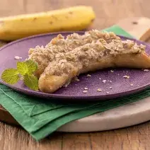 Foto da receita de banana grelhada recheada servida em um prato roxo sobre um paninho verde em uma tábua de madeira de forma circular. Ao fundo há uma banana para decoração