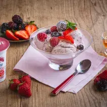 Fotografia em tons de rosa em uma bancada de madeira clara, um pote de vidro com o sorvete de frutas vermelhas, ao lado um potinho com mel e um pratinho com frutas vermelhas.