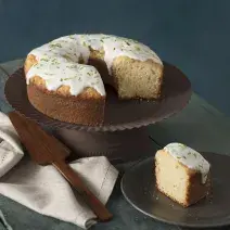 foto em tons de cinza, em uma mesa de madeira contém o bolo de iogurte ao lado um prato marrom com um pedaço de bolo um pano branco e por cima do pano uma espátula.