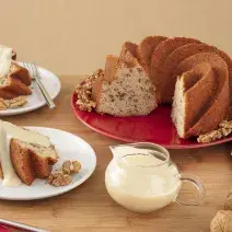 Foto da Receita de Bolo de Nozes com Molho doce. Observa-se dois pratos de sobremesa com pedaços do bolo e a calda. Ao lado direito, um prato redondo vermelho grande com o restante do bolo, decorado por nozes.