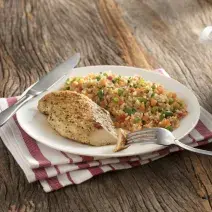 Fotografia em tons de marrom com um pano branco com listras vinho e um prato branco raso com o arroz feito na panela de pressão com legumes e um filé de frango.