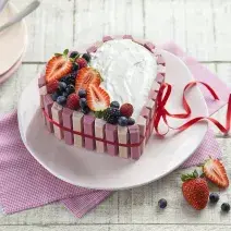 fotografia em tons de branco e rosa de uma bancada branca vista de cima, ao centro um pano rosa com um prato redondo por cima um bolo em formato de coração com marshmallow, morangos e frutas vermelhas por cima para decorar.