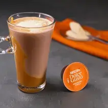 Fotografia em tons de laranja em uma bancada de madeira cinza escura, um paninho laranja e uma xícara de vidro alta com o café de mochaccino canela com doce de leite e achocolatado Galak.