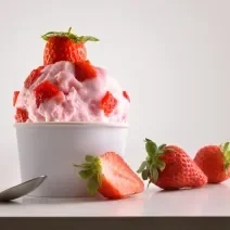 Fotografia de frozen iogurte de morango em um potinho de papel com pedaços de morango, e em cima, um morango inteiro. Ao lado, dois morangos inteiros e um morango cortado pela metade, no outro lado, uma colher de sobremesa, sobre um fundo branco.