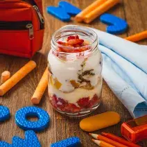 Fotografia em tons de laranja e azul em uma bancada de madeira escura, objetos escolares, paninho azul e ao centro, um potinho de vidro com o iogurte, Mucilon e frutinhas.
