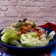 Foto da receita de Salada de Grão de Bico. Observa-se um prato azul redondo fundo com a salada com tomate, grão de bico, atum e alface americana dentro. Ao fundo, uma parede de tijolo branco.