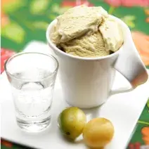Fotografia em tons de verde em uma mesa de madeira com uma toalha verde florida, um prato quadrado branco ao centro com uma xícara branca com o sorvete de umbu dentro. Ao lado, um copinho de vidro com água com gás e duas frutas de umbu.