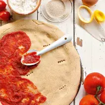 Fotografia em tons de vermelho e branco, de uma bancada de madeira com uma massa redonda de pizza, com molho de tomate pela metade e uma colher prateada. Ao redor potinhos, ovos, tomates e azeite.