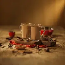 Fotografia em tons de marrom em uma bancada de madeira de cor marrom. Ao centro, um pano vermelho contendo 2 xícaras com o chocolate quente e ao redor há alguns pedações de chocolates espalhados.