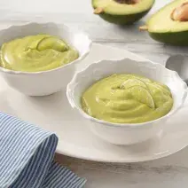 Fotografia em tons de branco e verde de uma bancada, ao centro dois potinhos com creme verde de abacate. Ao fundo, um abacate cortado ao meio.