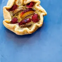fotografia em tons de azul e bege de uma bancada azul vista de cima, contém uma torta aberta com pedaços de framboesas frescas, pera e pêssegos