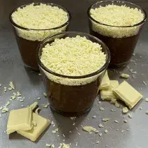 Foto da receita de trufa cremosa servida em três porções em copos de vidro baixos sobre uma mesa acinzentada com pedaços e raspas de chocolate branco rodeando os copos como decoração