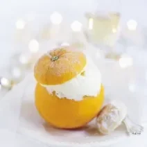 Fotografia em tons de branco com uma laranja inteira recheada de creme apoiada sobre um prato, em cima de uma mesa branca. Ao fundo, uma taça com champanhe.