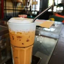 Fotografia de uma bebida de morango e maracujá com leite condensado e gelo que sobre um apoio pequeno de cor preta. Dentro do copo tem um canudo preto, e a bebida está sobre uma mesa escura com vidro.