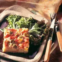 Fotografia em tons de verde em uma bancada de madeira com uma toalha bege colorida, um prato quadrado roxo com o pedaço da torta com azeitonas e ovos e ao lado salada de folhas verdes.