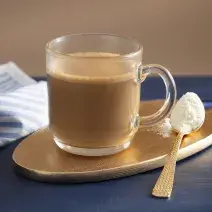 Fotografia em tons de marrom em uma bancada de madeira de cor azul. Ao centro, uma tábua de madeira dourada contendo uma xícara de café com uma colher de leite em pó ao lado. Ao fundo, há um pano listrado azul.