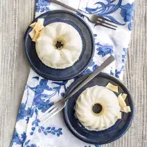 Fotografia em tons de azul em uma bancada de madeira clara, um pano branco com folhas azuis e dois pratinhos azuis escuros com o mini pudim de tapioca feito com iogurte, abacaxi e coco.