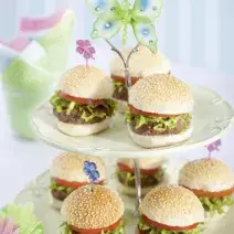 Fotografia em tons de verde em uma mesa branca com um pedestal enfeitado com borboletas de papel de pratos brancos com os hamburguinhos colocados nele. Ao fundo, um copo com guardanapos de papel verde e rosa.