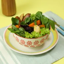 Fotografia em tons de amarelo em uma bancada de madeira amarela, um jogo americano verde claro listrado, um prato branco com listas coloridas, um potinho branco com flores e dentro uma salada de legumes.