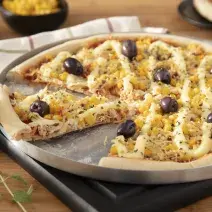 Fotografia em tons amarelo em uma bancada de madeira, uma tábua de alumínio redonda com a pizza de frango com catupiry em cima dela. Ao lado, potinhos com milho, azeitonas e um prato com uma fatia da pizza.