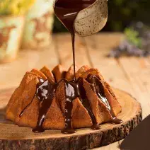 Fotografia em tons de marrom em uma bancada de madeira e ao centro uma tábua de tronco de árvore com o bolo de cenoura em cima e uma mão segurando um potinho despejando a calda de chocolate por cima do bolo.