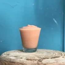 Fotografia de um frozen iogurte com goiaba dentro de um copo de vidro sobre um apoio de madeira, e ao fundo, uma parede na cor azul.