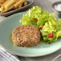 Fotografia em tons de verde em uma bancada de madeira clara, um pano azul, um prato verde claro com o steak tartare acompanhado de salada de alface e tomate cereja. Ao fundo, um recipiente com uma porção de batatas fritas.