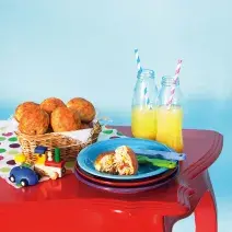 Fotografia em tons de azul e vermelho em uma mesa antiga vermelha com pratos coloridos, cesta de vime com os lanchinhos de frango sem leite e sem ovo e garrafas de vidro sobre a mesa.