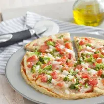 fotografia em tons de branco de uma bancada vista de frente, contém um prato redondo e branco com uma pizza com pedaços de tomates e brócolis ao fundo uma jarra com azeite.