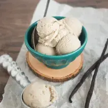Fotografia em tons de verde em uma bancada de madeira com um pano branco amassado, um pote verde com o sorvete de baunilha e mel dentro. Ao lado, um pegador de sorvete com uma bola dentro dele.