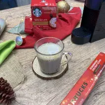 Fotografia em tons de vermelho em uma bancada de madeira com um pano vermelho, uma xícara de vidro com o café e enfeites natalinos.