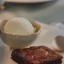 Foto da receita de Bolo Quente com Geleia da consumidora Caroline Lima. Observa-se um bolo baixo de chocolate com calda brilhosa por cima e, atrás, em um pote de cerâmica, uma bola de sorvete de creme.