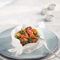 Fotografia em tons de azul em uma mesa com uma toalha branca de renda, um prato branco e azul claro em cima dela e o papillote de legumes (abobrinha, cenoura, tomate) em cima do prato azul, enrolado em um papel manteiga.