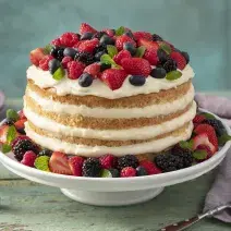 Fotografia em tons de vermelho e lilás em uma bancada de madeira verde, um pano roxo, um suporte para bolo com o bolo pelado (naked cake) com creme branco e com decoração de frutas vermelhas. Ao fundo, pratos pequenos lilás de sobremesa.