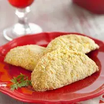 Fotografia em tons de vermelho em uma bancada de madeira com um pano branco e detalhes vermelhos, um prato vermelho redondo raso com três pasteis assados de frango. Ao fundo, uma taça vermelha com água.