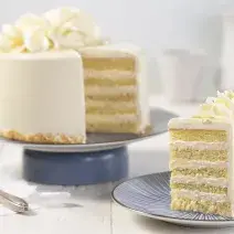 Fotografia em tons de branco, em uma bancada de madeira de cor branca. Ao centro, uma boleira azul contendo o bolo. Ao lado um pires contendo uma fatia de bolo e ao fundo uma jarra branca.