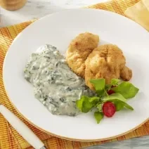 Foto da receita de Creme de Espinafre Sem Lactose. Observa-se um prato branco com o creme do lado esquerdo e, ao lado direito, uma posta de peixe frito com salada.