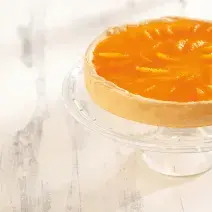 foto em tons de branco e laranja de uma bancada branca vista de cima. Contém um suporte para servir sobremesas transparente com uma torta com cobertura de tangerina por cima.