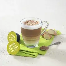 Fotografia em tons de verde em uma bancada de madeira clara com um pano verde e uma xícara de vidro com o cappuccino dentro dele. Ao lado, cápsulas de Dolce Gusto Cappuccino.