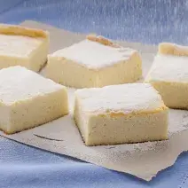 Fotografia em tons de amarelo, azul e branco de uma bancada branca com paninho azul, sobre ele pedaços de torta de ricota polvilhadas com açúcar.