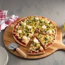 Fotografia em tons de vermelho em uma bancada de madeira cinza com uma tábua de pizza de madeira com a pizza em cima decorada com escarola e ricota. Ao lado, um vidro de azeite, tomates e um cortador de pizza.