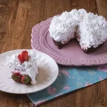 Imagem da receita de Torta Brownie, decorada com chantilly e morangos, servida em um prato rosa e ao lado há um prato menor branco com uma fatia. Tudo está sobre um pano decorado com motivos florais, sobre uma bancada de madeira