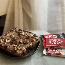 Foto aproximada de um Brownie de KitKat, em formato quadrado, sobre um prato preto. Ao lado dele há três KitKtas, um ao leite, outro dark e outro white.